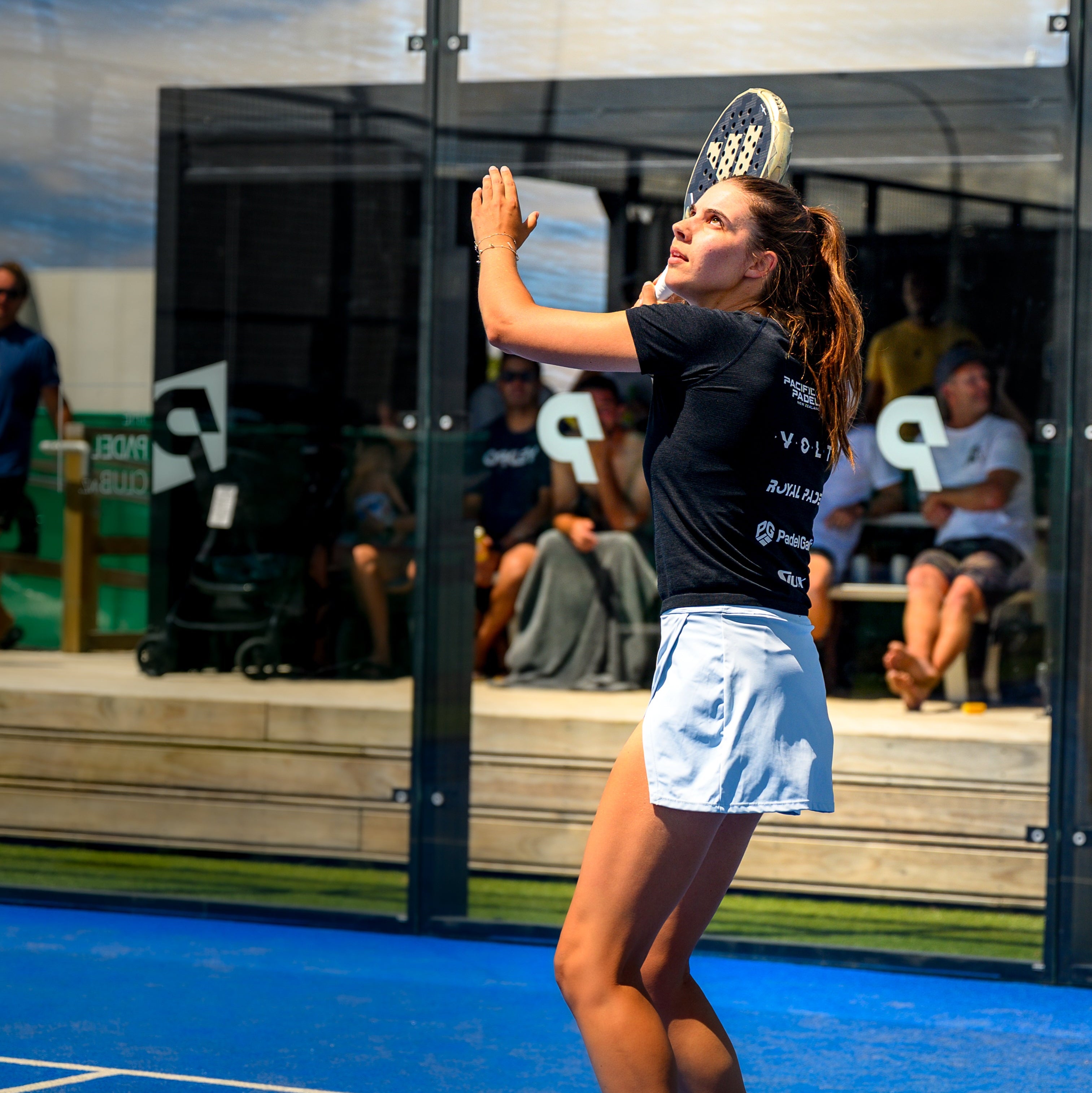 Woman playing paddle tennis on a blue court with spectators in the background.